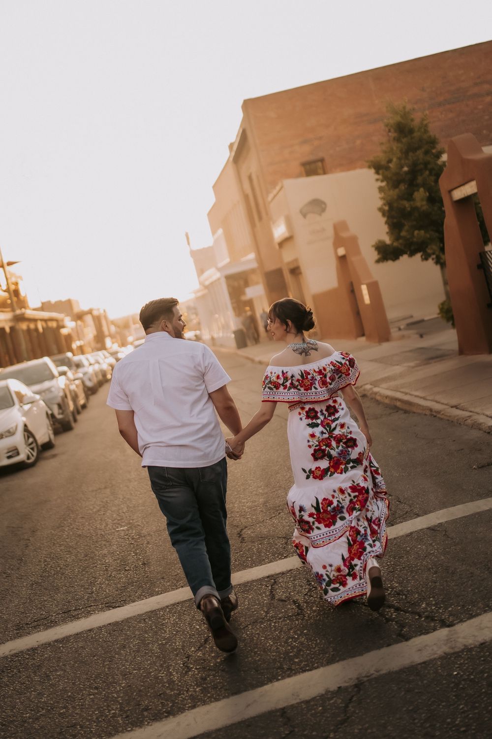 A couple walks down a sunset-lit street in Santa Fe New Mexico, with the woman wearing a red floral dress. Santa Fe wedding photographer.