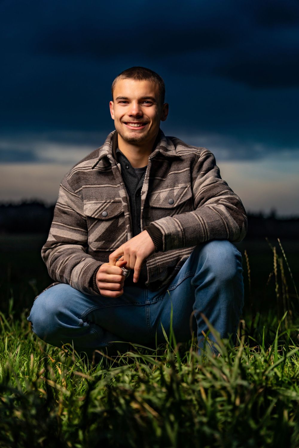A person in a brown leather jacket sits casually in green grass against a moody evening sky.