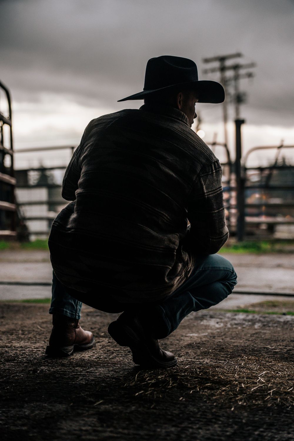 A person in cowboy boots crouches low to examine the dusty ground at a ranch corral during an overcast day.