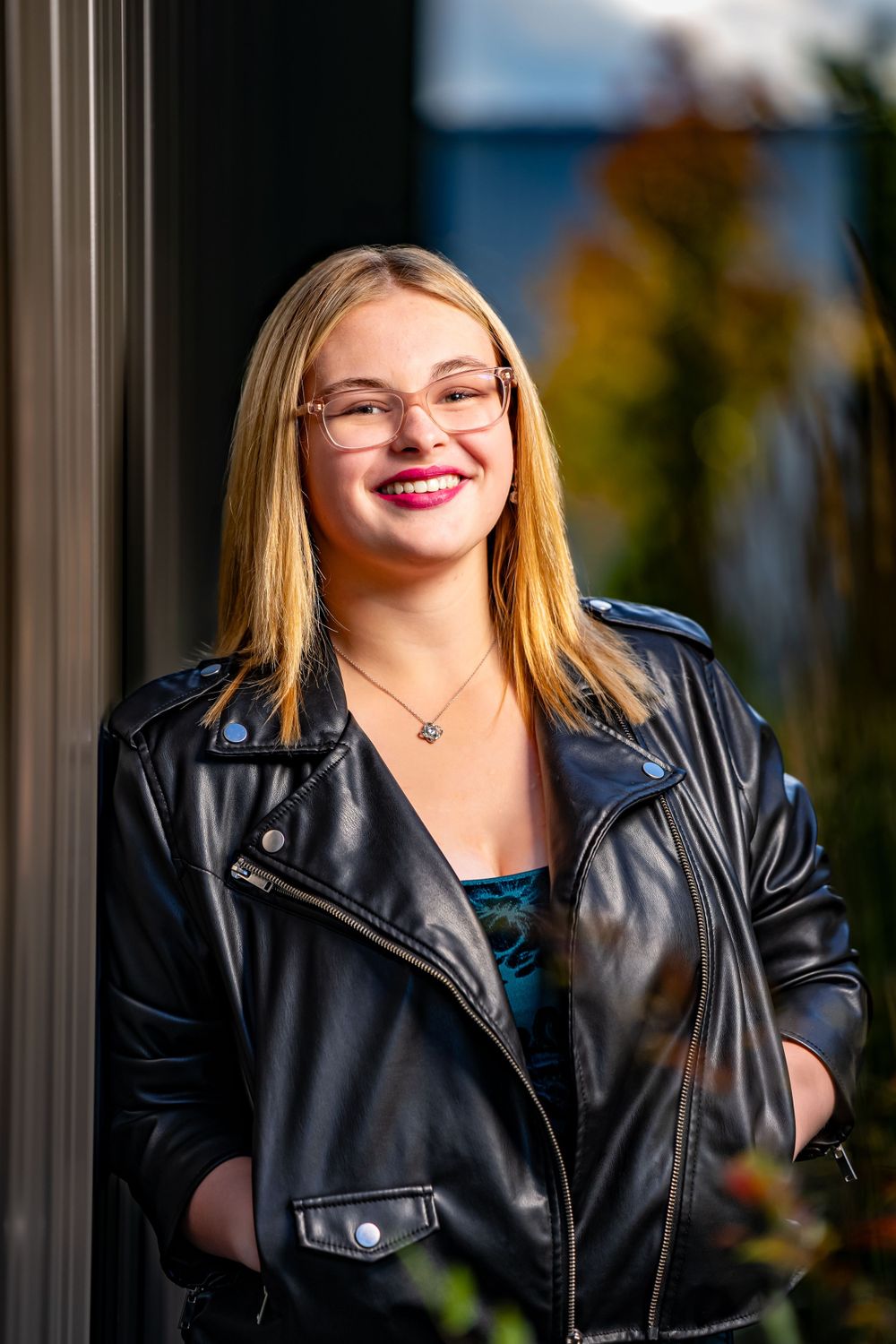 Stylish portrait of someone in a black leather jacket and glasses against a blurred outdoor background.