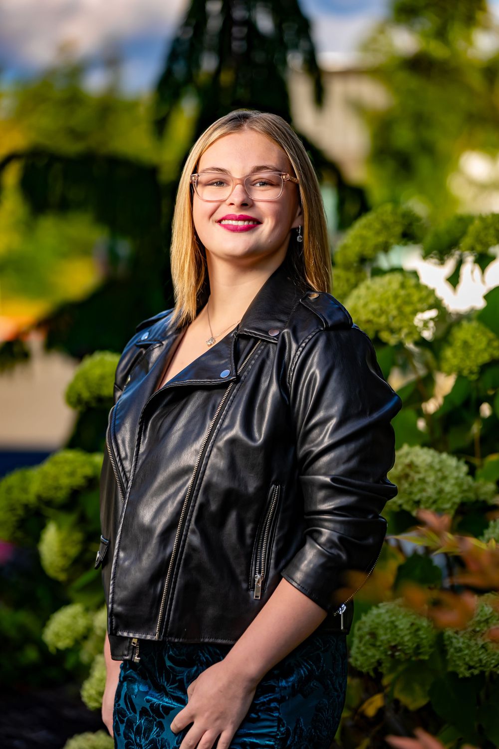 Casual outdoor portrait featuring a leather jacket and glasses with greenery in background.