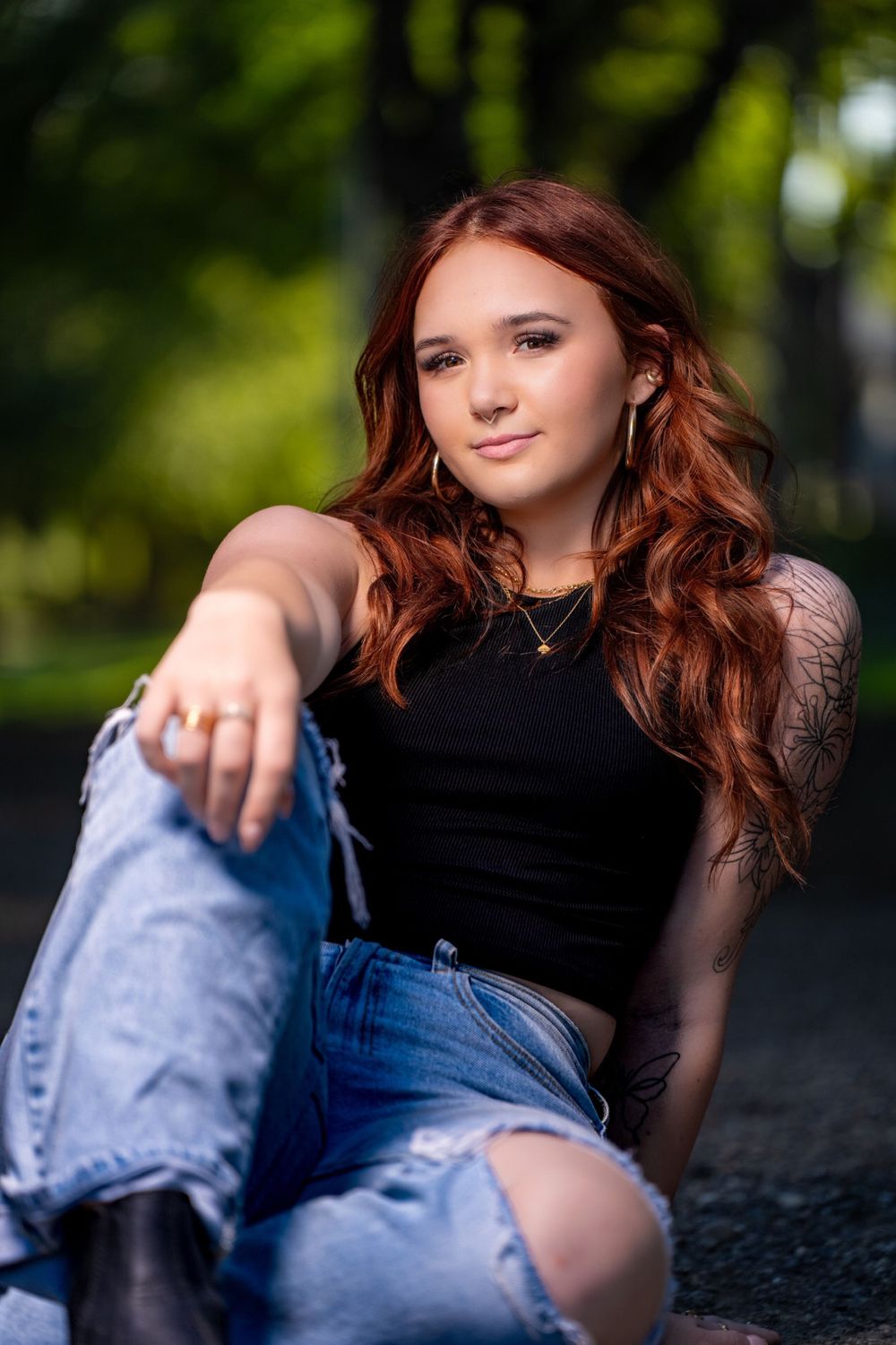 Portrait of someone in black top and ripped jeans seated casually outdoors with lush green background.
