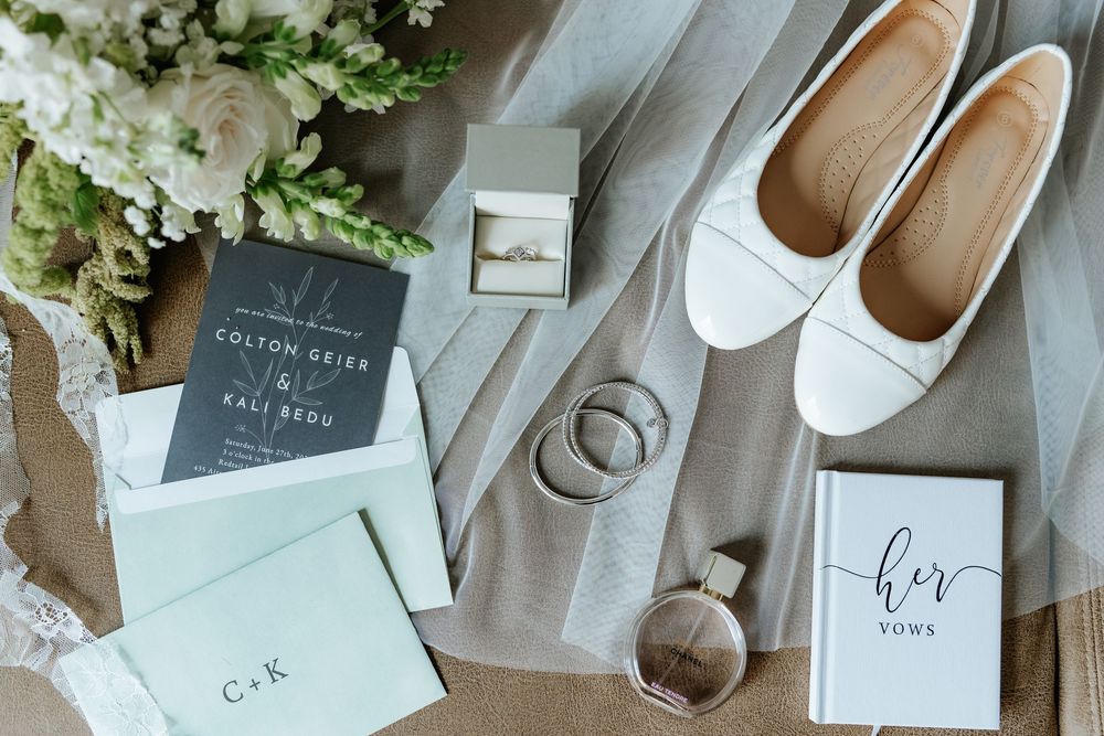Flat lay of wedding details including white flats, a ring box, bands, perfume, bridal bouquet, navy invitation suite and a her vows booklet.