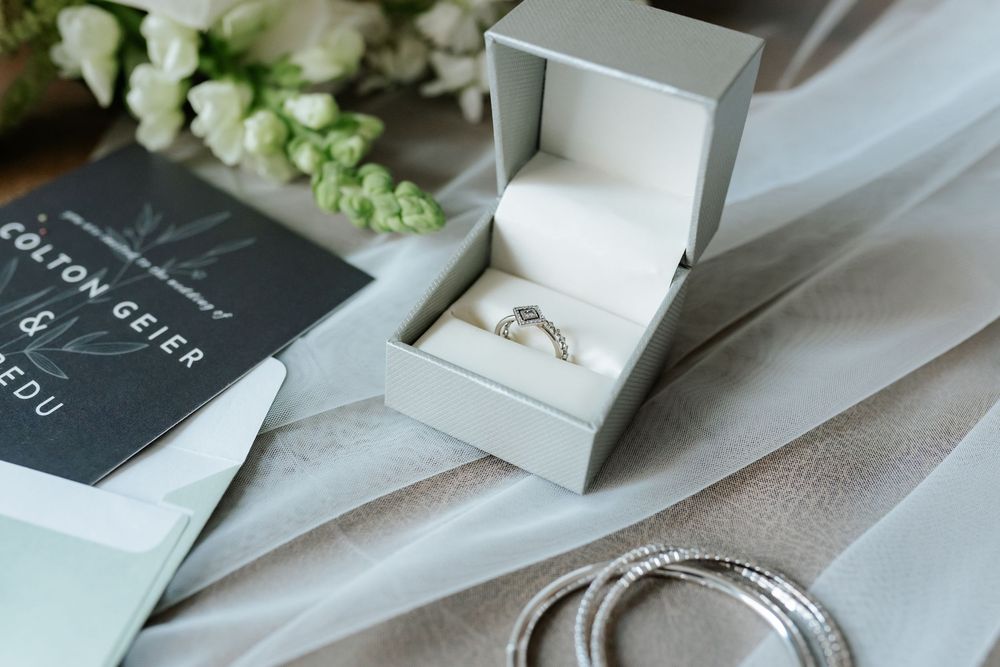 Diamond engagement ring in an open gray ring box beside a navy wedding invitation, white flowers, and silver bracelets on soft tulle.