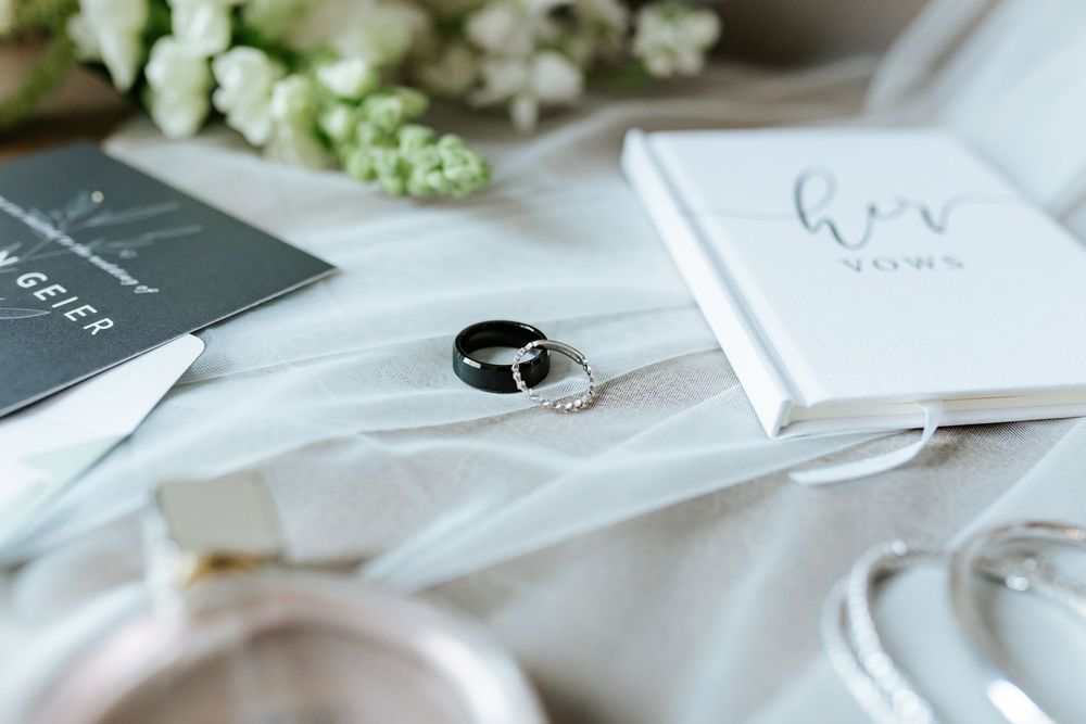 Black and silver wedding rings rest on soft white tulle beside a vow book, white flowers, and a dark invitation card.
