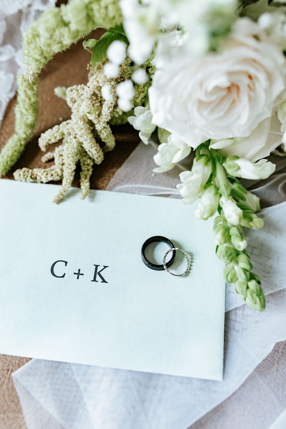 Wedding rings rest on a card marked C + K, beside a white rose and green bridal bouquet with delicate blooms.
