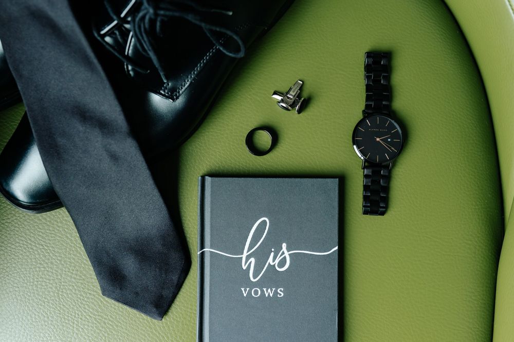 Groom's wedding morning flat lay with black tie, black watch, cufflinks, ring and a his vows booklet on a green chair.