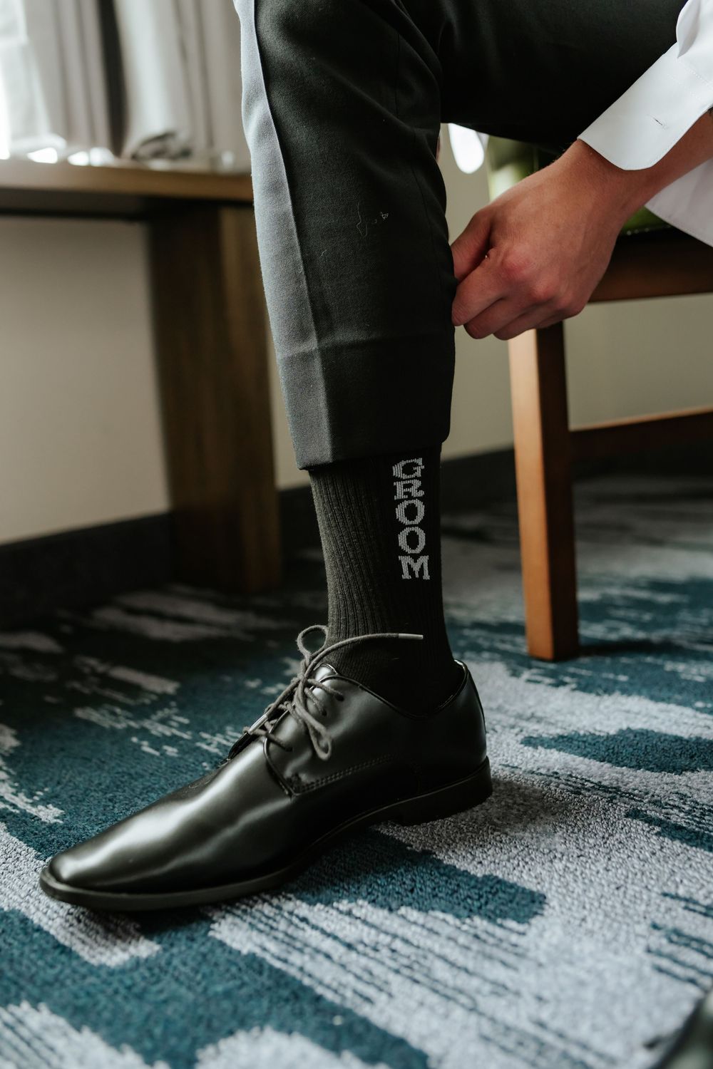 Groom sitting in a hotel room wearing polished black leather dress shoes and dark socks that read GROOM on a patterned teal carpet.