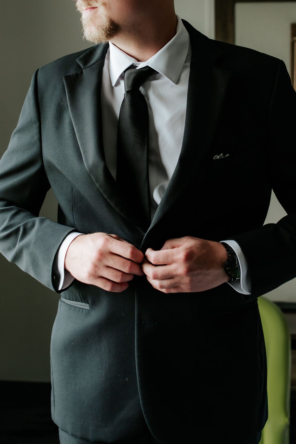 Groom in a charcoal suit and black tie buttons his jacket while getting ready for his wedding day.
