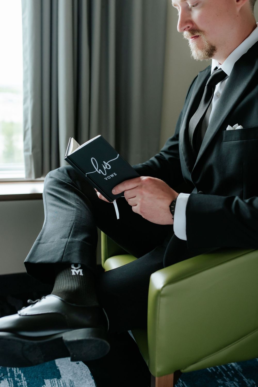 Groom in a dark suit sits in a green armchair by a window, reading his handwritten wedding vows booklet.