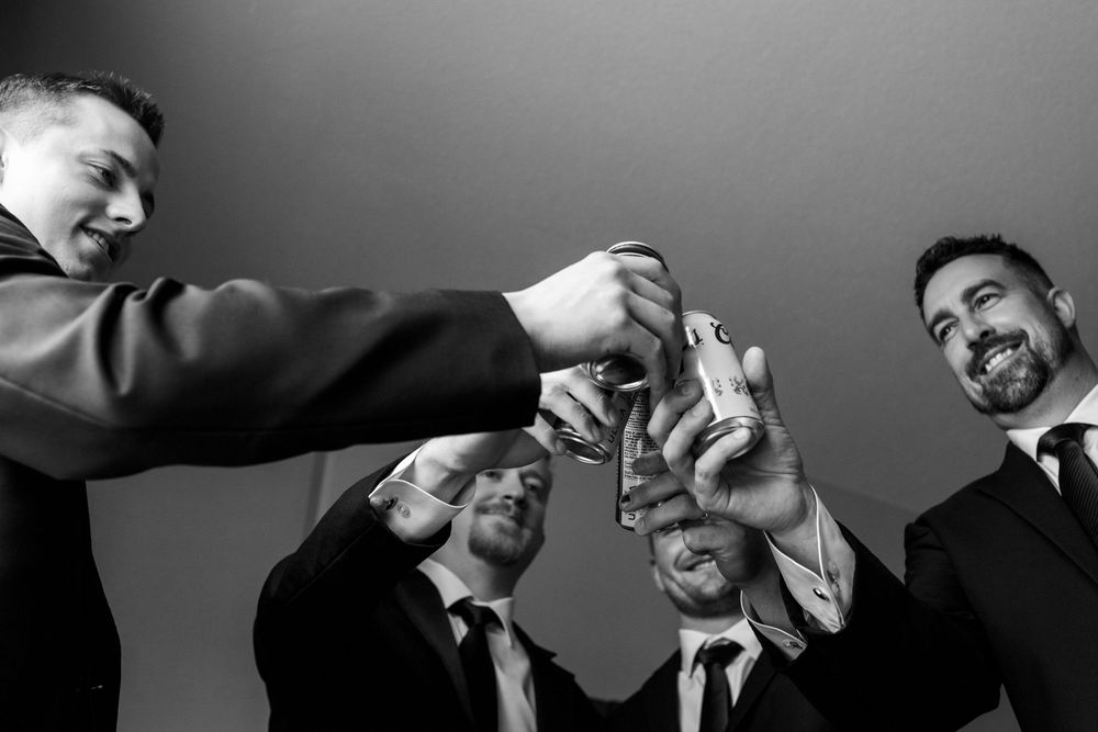 Four groomsmen in suits and ties lean in close to clink drink cans together in a joyful black and white toast before the wedding.