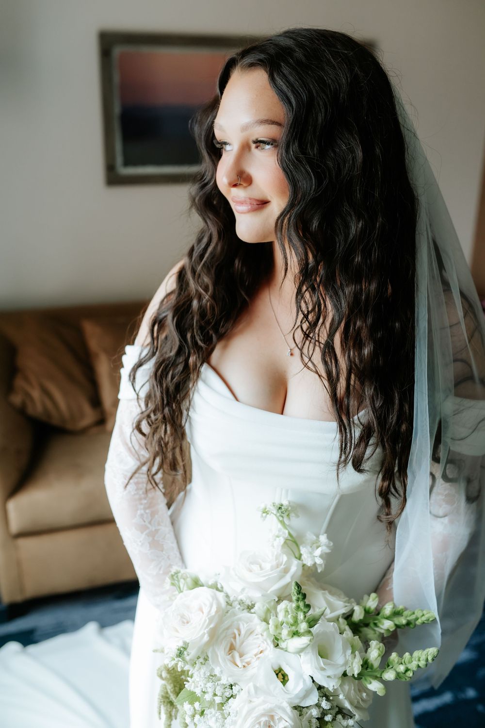Smiling bride with long wavy dark hair, lace sleeves and a veil holds a white rose bouquet while gazing out a window.