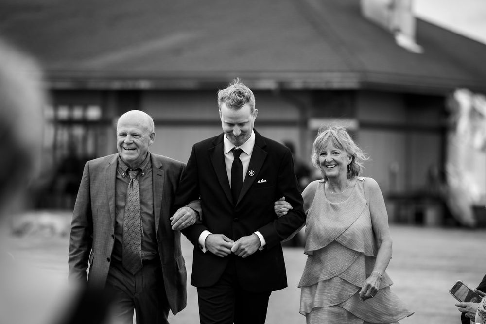 Smiling groom in a dark suit walks arm in arm with a man and a woman in a black and white wedding photo.