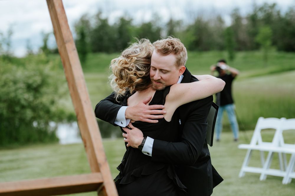 A woman with curly blonde hair hugs a smiling man in a black suit at an outdoor wedding with a wooden arch and white chairs.
