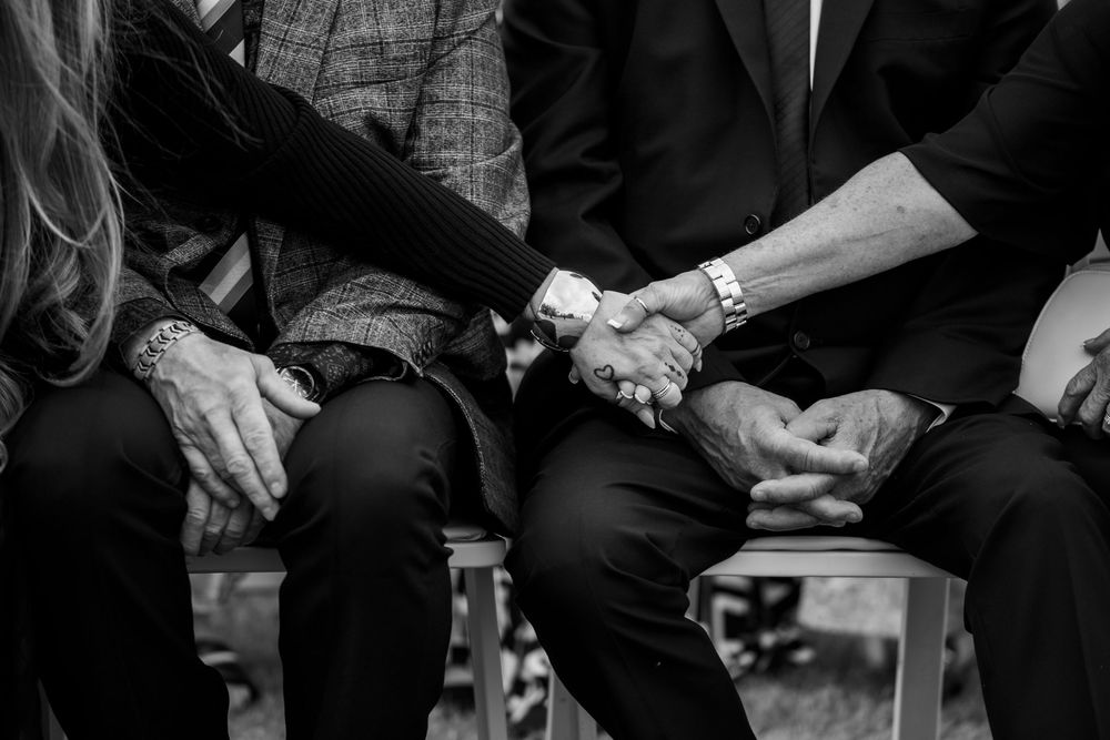 Black and white close-up of seated guests holding hands in comfort and support during an emotional wedding ceremony moment.