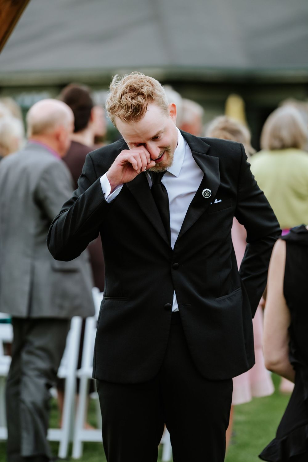 Groom in a black suit and tie wipes a tear from his eye while standing on a lawn at an outdoor wedding ceremony with guests behind him.