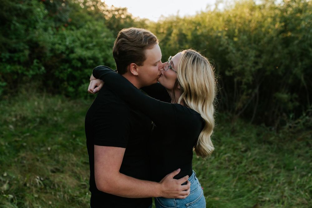 A couple in black shirts shares a romantic kiss while embracing in a lush green field surrounded by trees and shrubs at sunset.