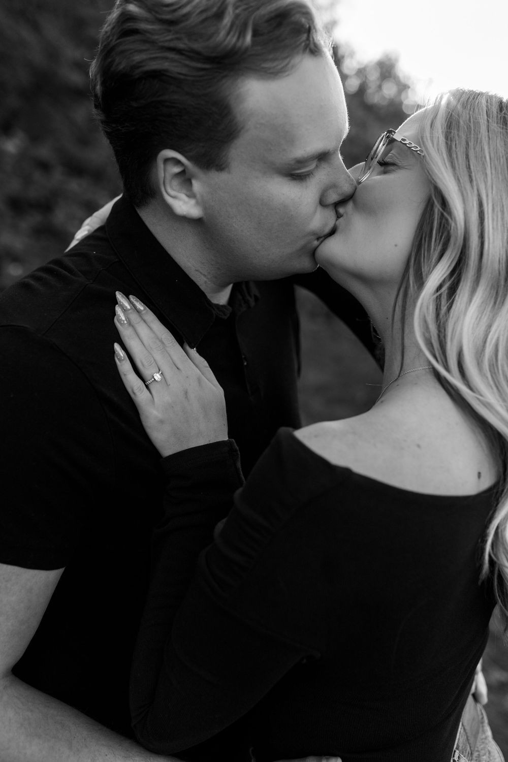 Black and white photo of a couple kissing outdoors, the woman's hand on his shoulder showing a sparkling engagement ring.