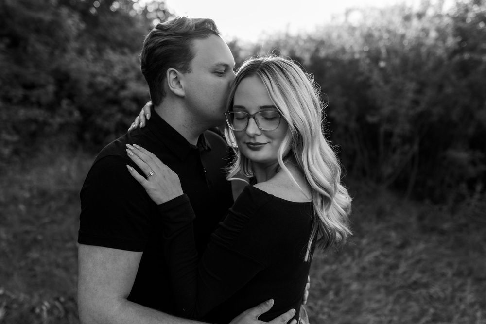 Black and white photo of a couple embracing outdoors as a man kisses the forehead of a smiling woman with long wavy hair and glasses.
