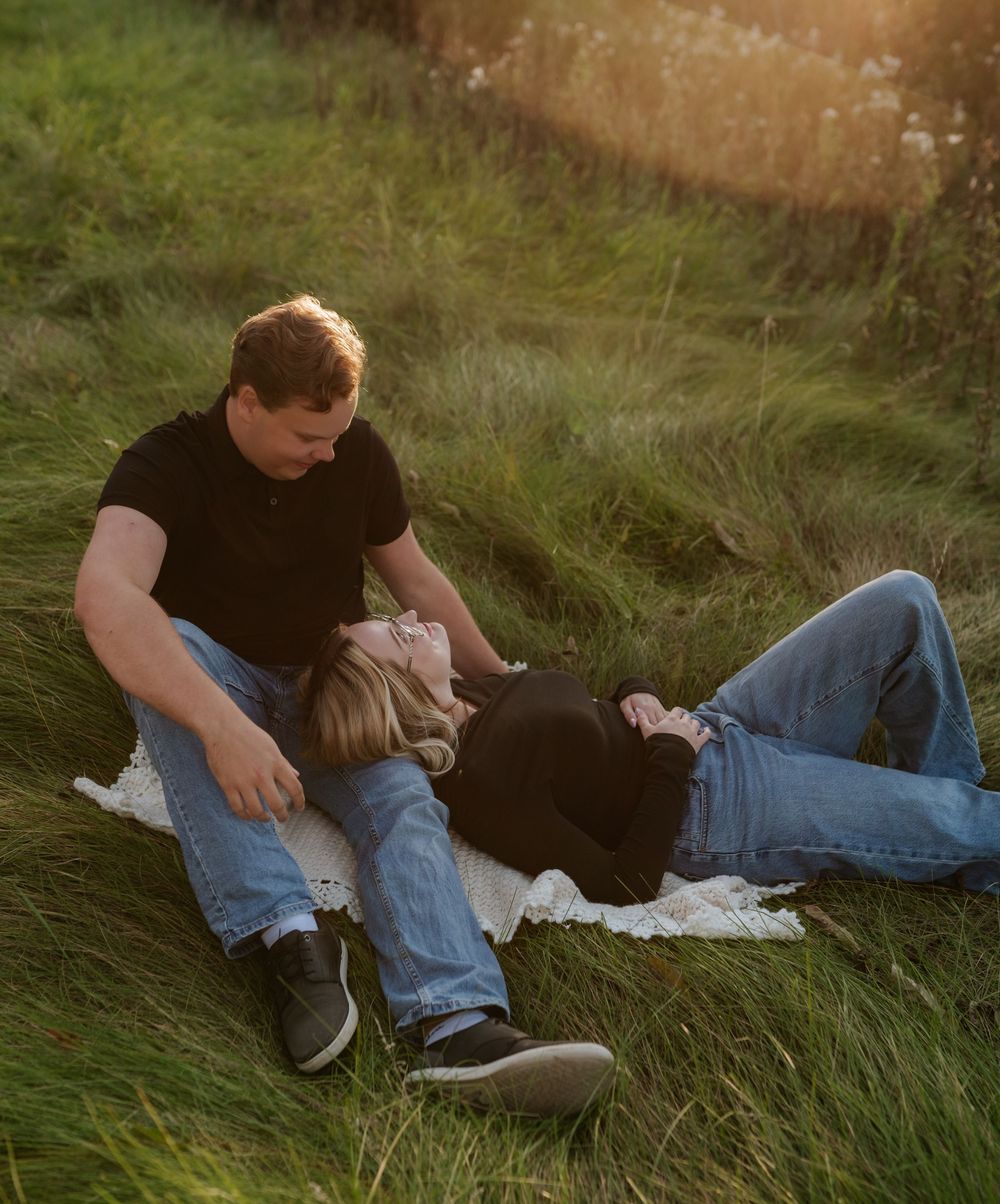 Couple relaxing together on a white blanket in tall green grass, bathed in warm golden sunset light.
