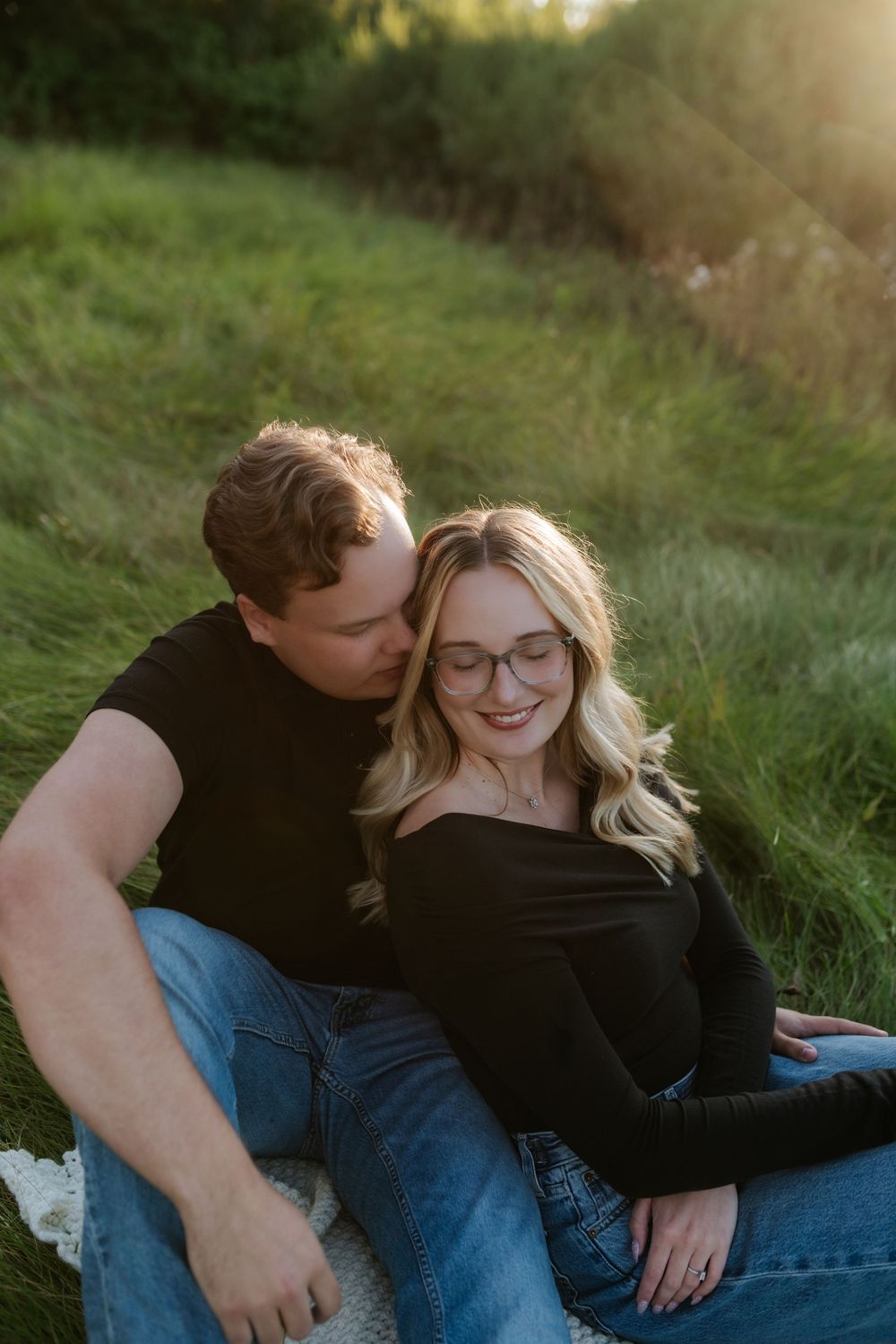 Smiling woman with glasses and long blonde hair snuggles with a man in a black shirt while sitting on a grassy hillside at golden hour.