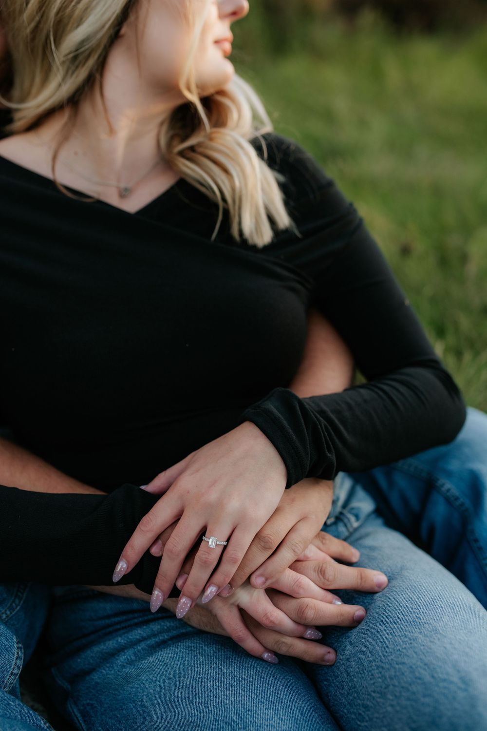 Close-up of a couple sitting on the grass with hands clasped, showing a sparkling diamond engagement ring and manicured nails.