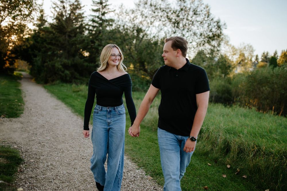 Smiling couple holding hands and walking along a gravel path beside a grassy meadow and trees during an outdoor engagement photo session.