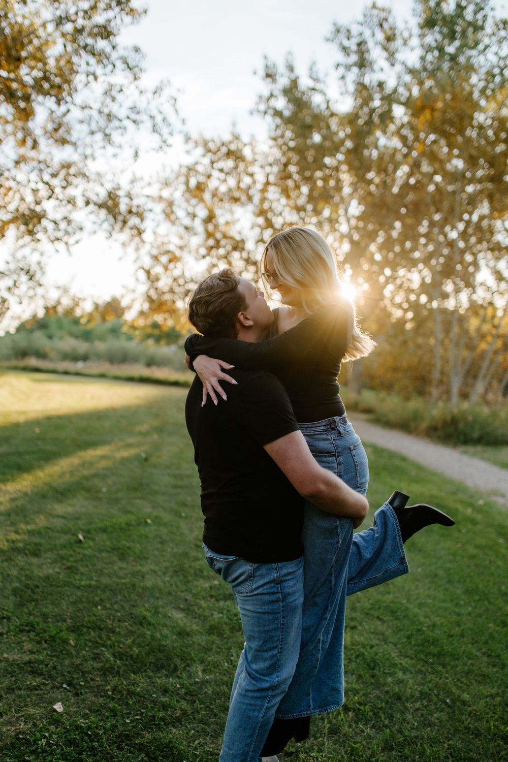 Smiling man in a black t-shirt lifts his blonde partner in an embrace on a grassy park lawn during a sunset engagement photo session.