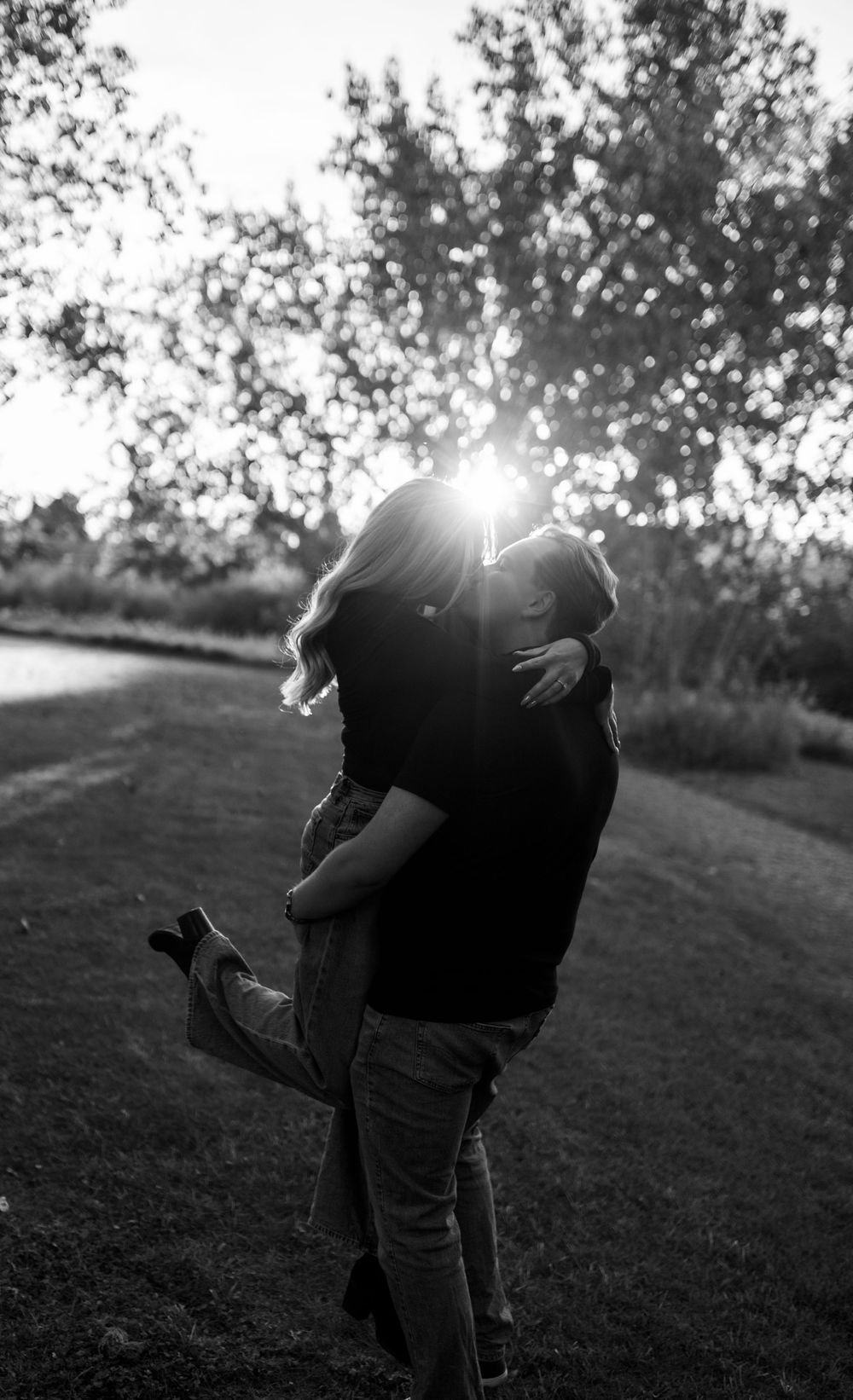 Black and white photo of a couple embracing in a sunlit park, the woman lifted with one foot raised as the sun glows through the trees.