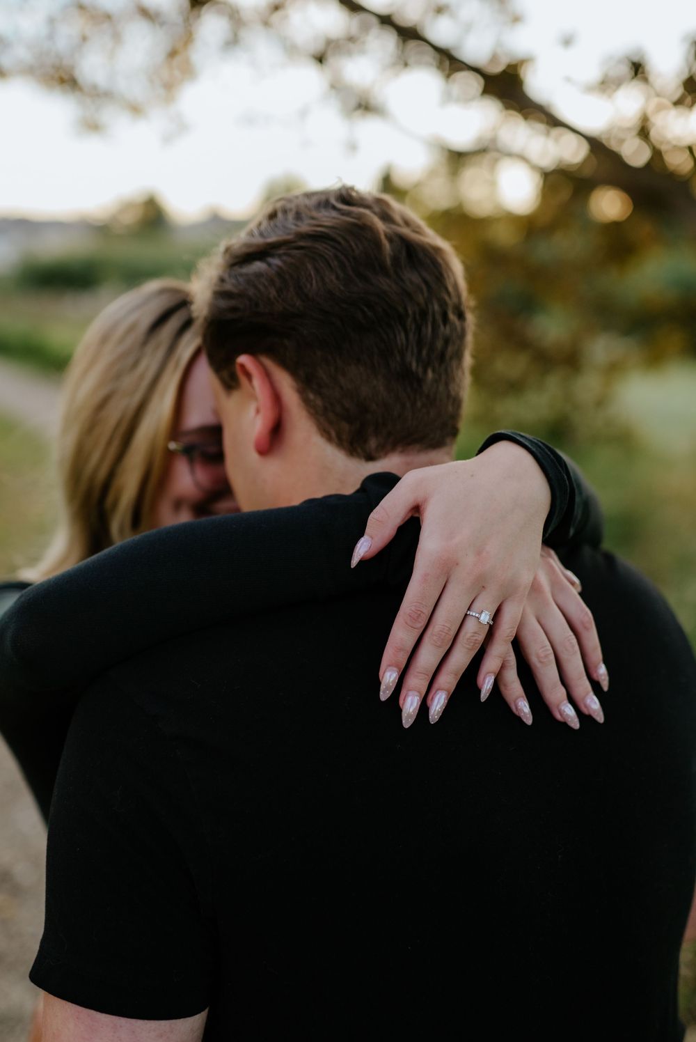 Couple embracing outdoors at sunset as the woman wraps her arms around his neck, her engagement ring and manicured nails in focus.