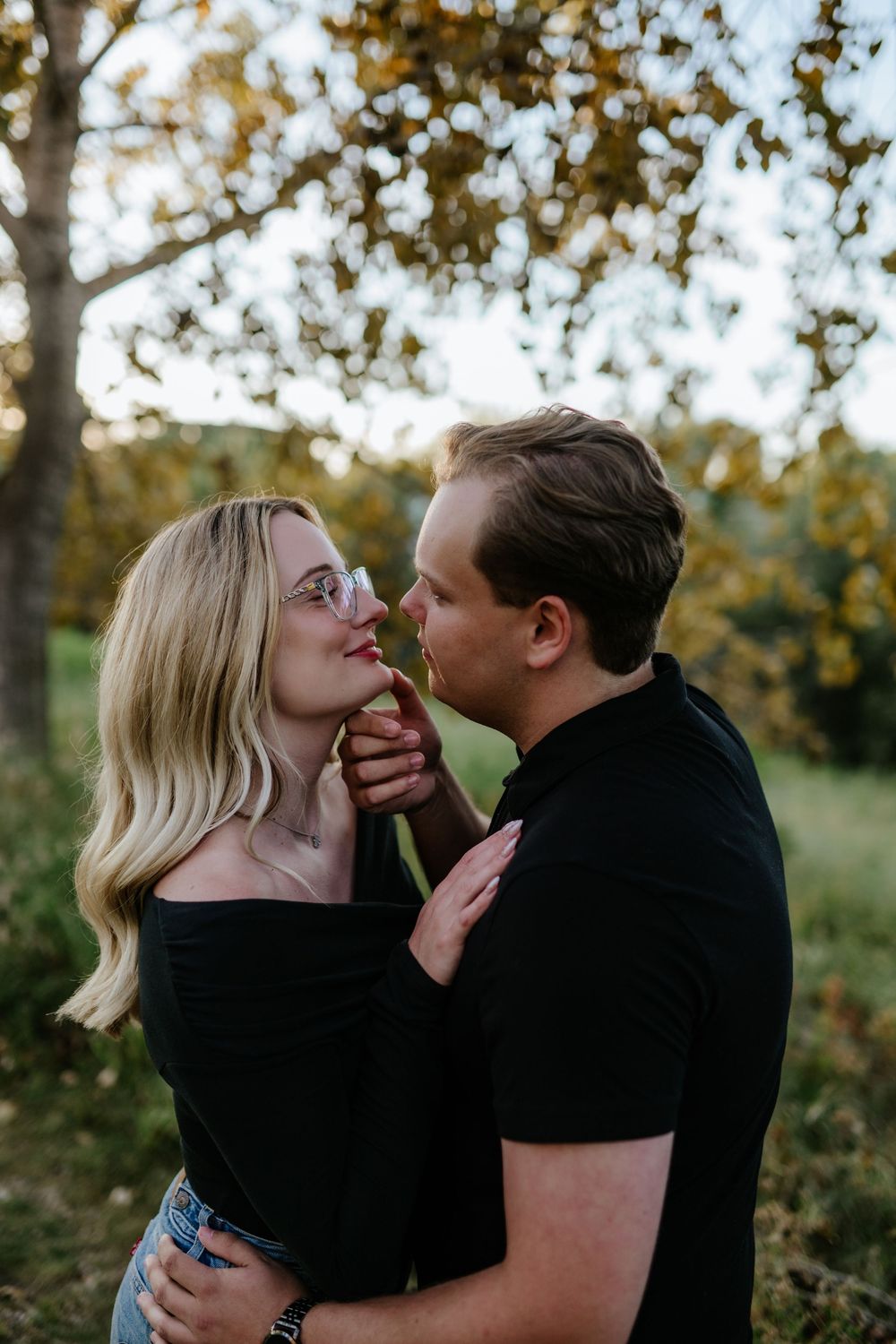 Blonde woman in glasses and black top touches her partner's chin as the couple share an intimate moment among autumn trees at golden hour.