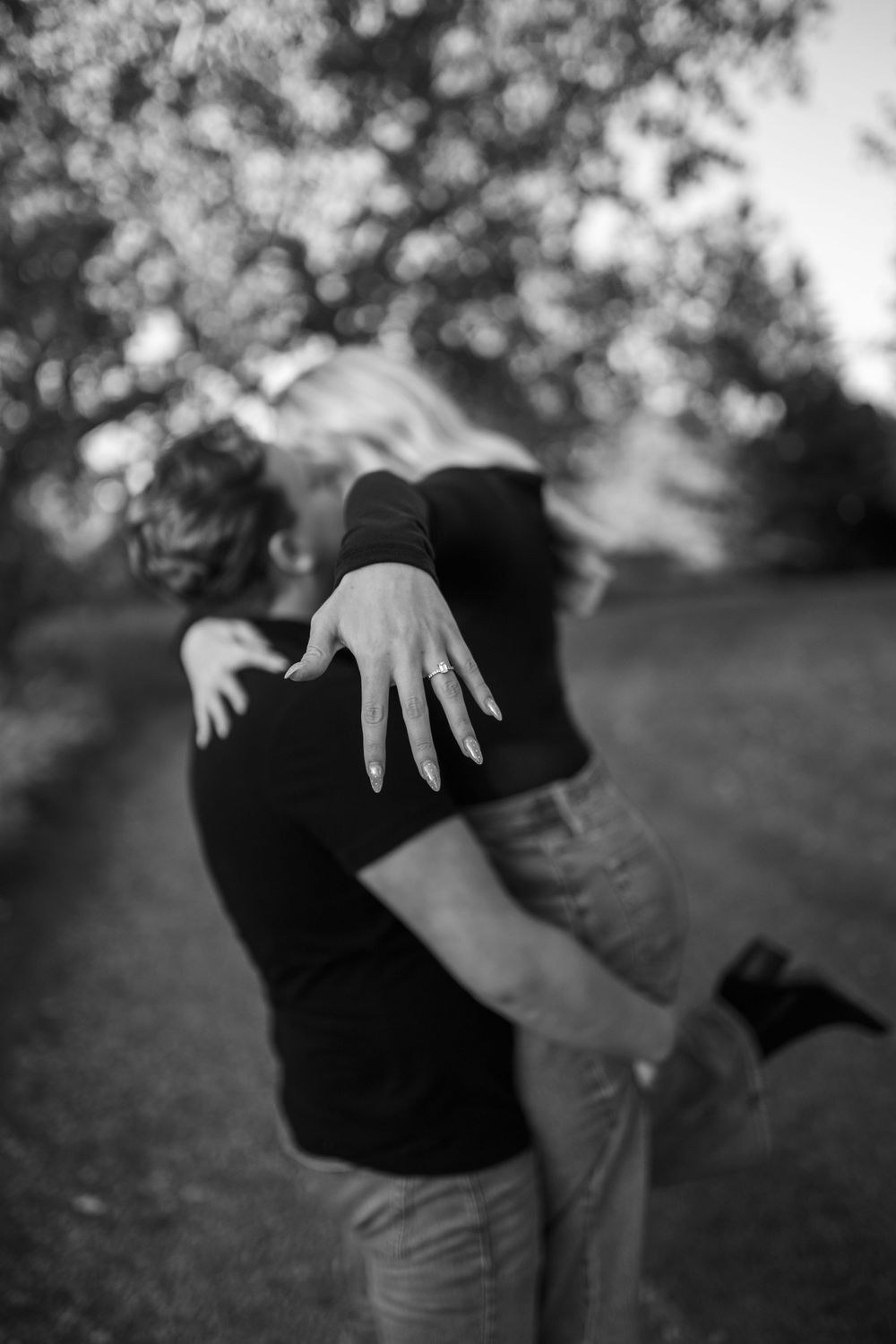 Black and white engagement photo of a couple embracing outdoors as the woman extends her hand to show off her ring among blooming trees.