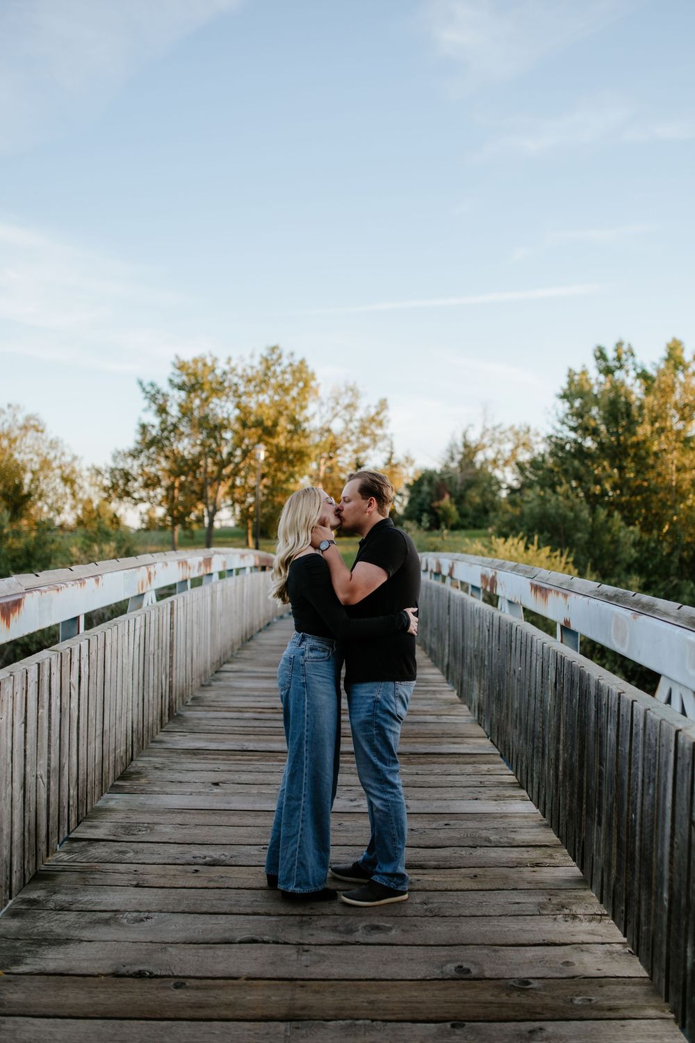 Couple in jeans and black tops embrace and kiss on a wooden bridge surrounded by green trees under a soft blue sky at golden hour.