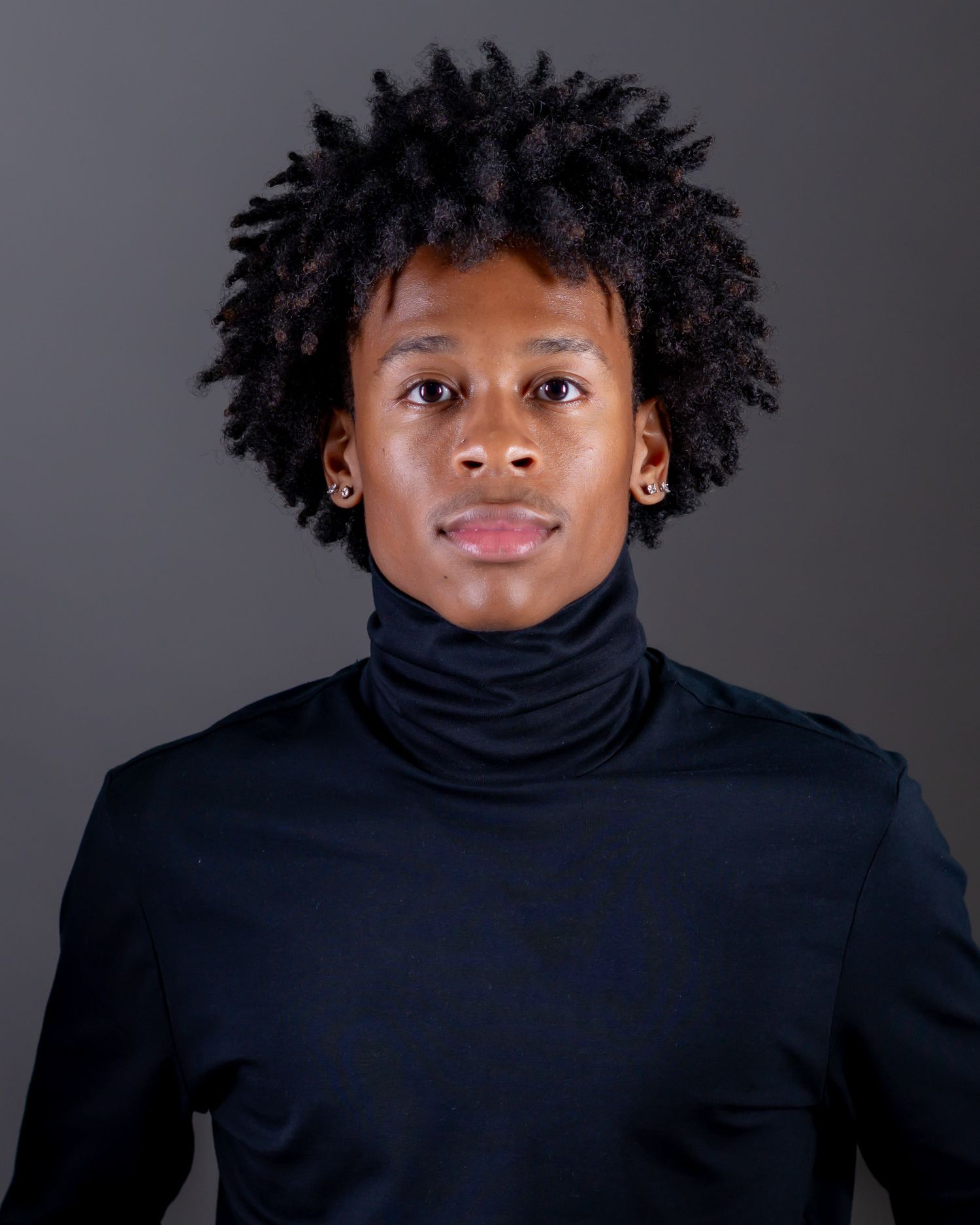 Portrait of a young man with short curly black hair, small stud earrings, and a black turtleneck against a gray studio background.