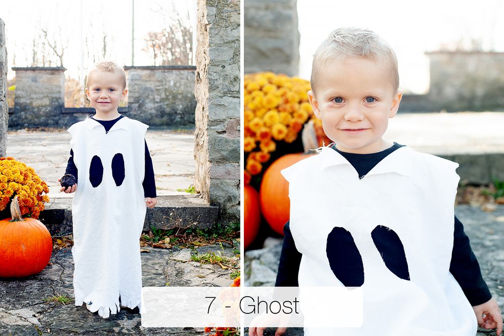 Young boy wearing a homemade white ghost costume with black oval eyes, standing beside orange pumpkins and yellow mums on a stone patio.