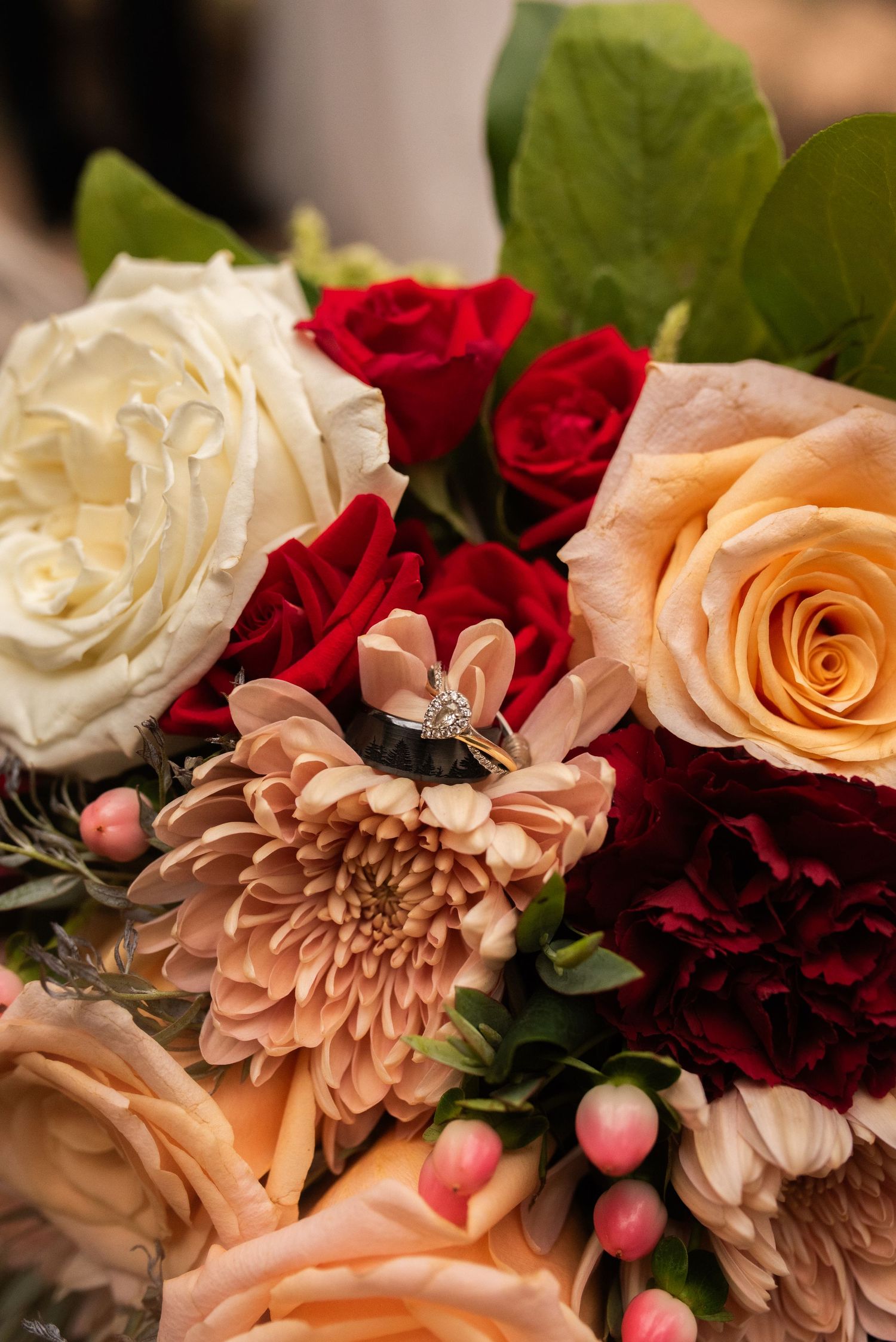 Diamond engagement ring resting on a wedding bouquet of cream and peach roses, red roses, burgundy carnations, and pink hypericum berries.