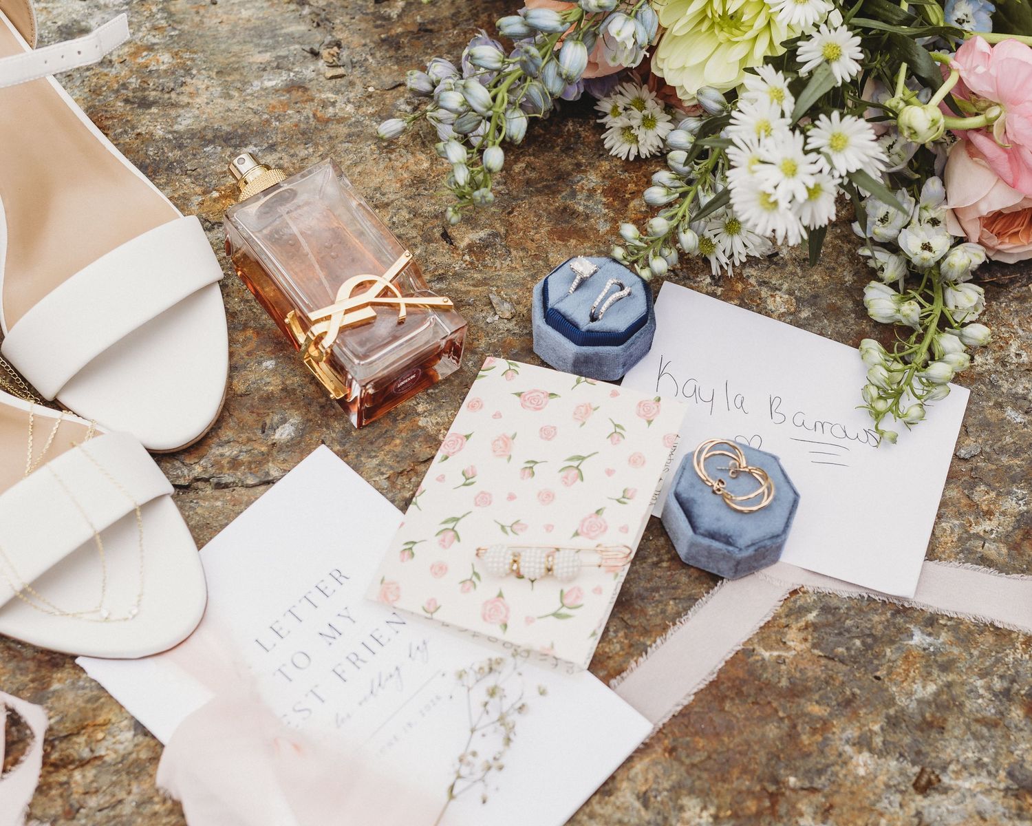 Wedding flat lay featuring white bridal shoes, perfume, blue velvet ring boxes, vow books, stationery, and flowers.