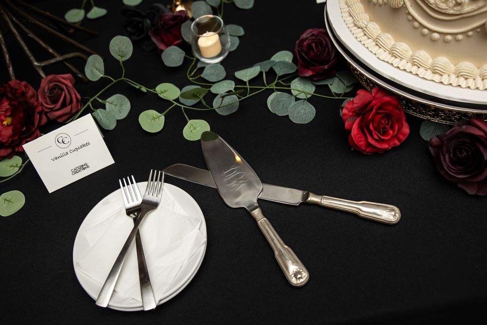 Wedding cake table with a silver cake server and knife, two forks on a white plate, red roses, eucalyptus garland, and a tiered cake.