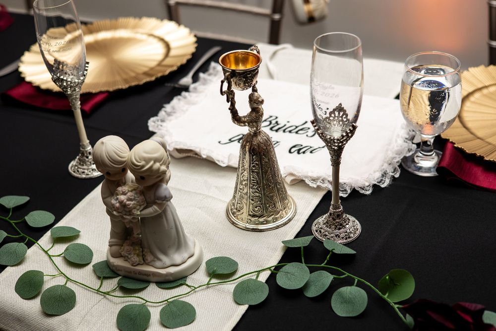 Wedding table setting with a bride and groom figurine, ornate goblets, wine glasses, gold chargers, and eucalyptus garland on black linen.