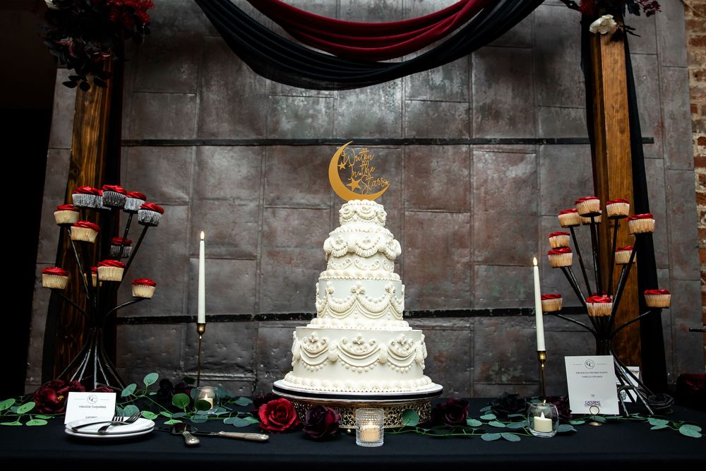 Tiered white wedding cake with a gold crescent moon topper on a dark gothic table with candles, red roses and a rustic metal backdrop.