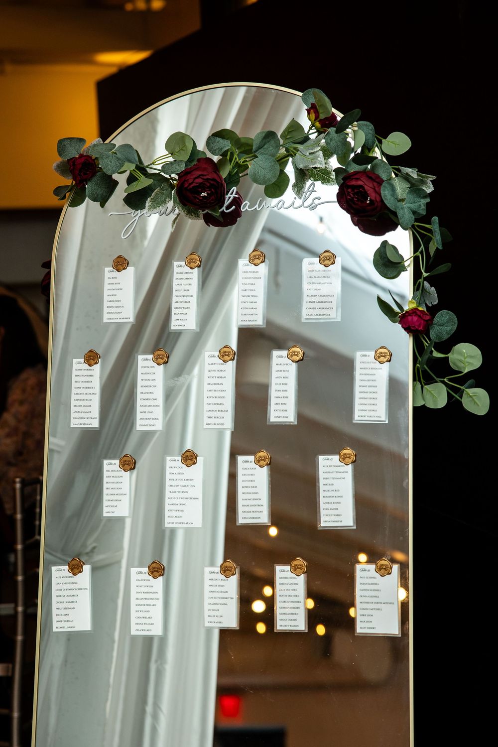Arched mirror wedding seating chart with gold wax seals, name cards, and burgundy roses with eucalyptus garland.
