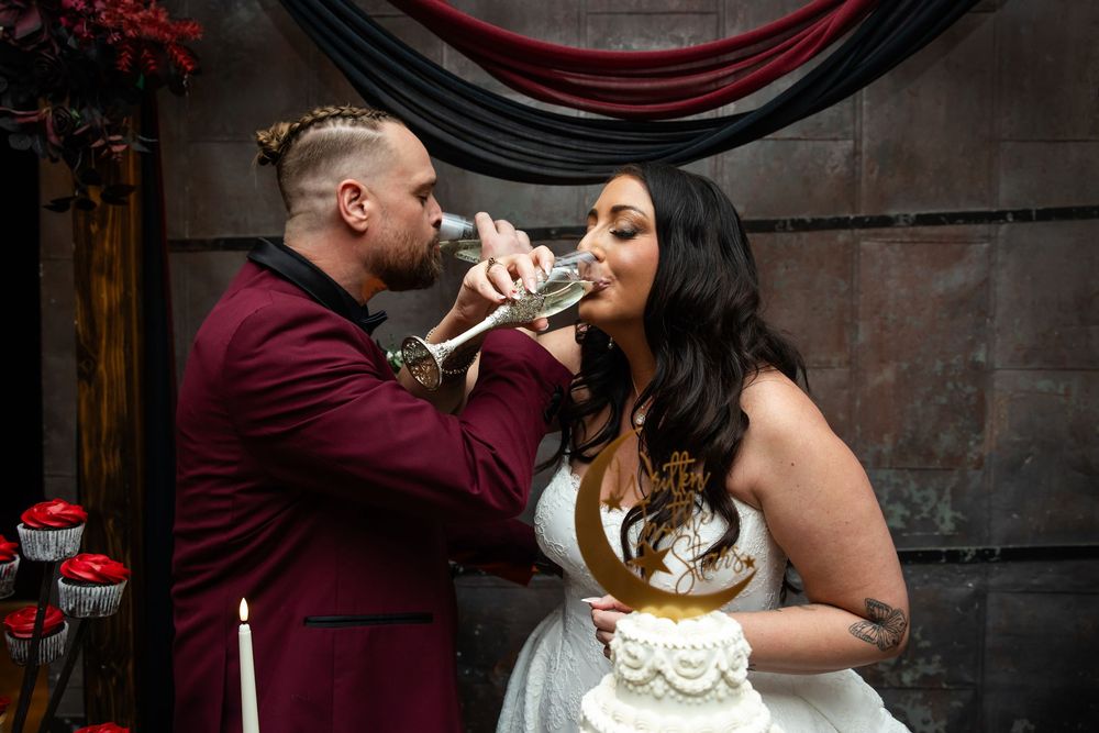 Bride in white dress and groom in burgundy velvet jacket toast with champagne flutes beside a wedding cake at a moody gothic reception.