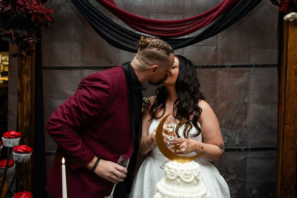 Newlywed couple kissing beside a white wedding cake, the groom in a burgundy suit and the bride in a white gown, holding champagne glasses.