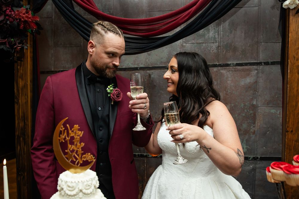 Bride in a white gown and groom in a burgundy blazer smile at each other while holding champagne flutes at their wedding reception.
