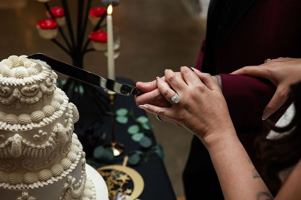 Newlyweds cut their ornate white tiered wedding cake together with a knife beside a lit candle and red floral decor.