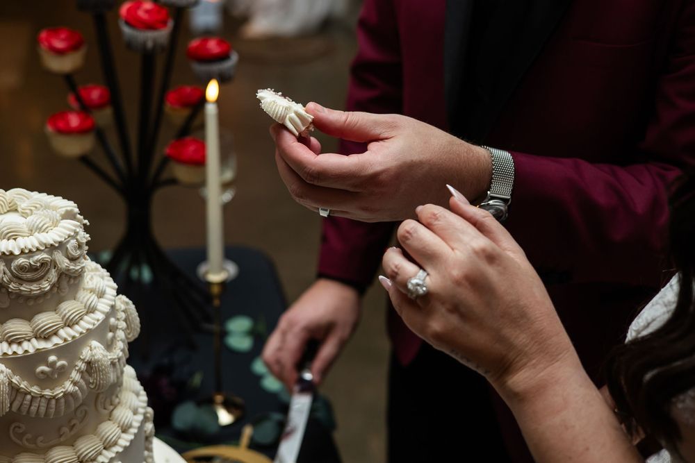 Couple in burgundy attire sharing a bite of wedding cake beside a lit candle, with a tiered white cake and red candle holders nearby.