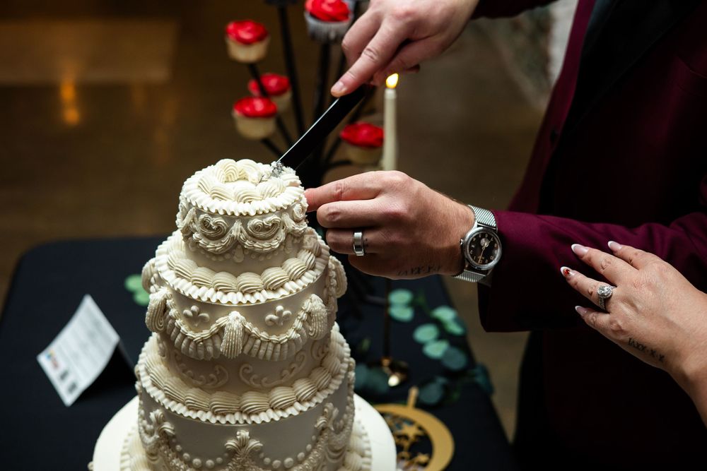 Couple cutting a vintage-style white tiered wedding cake with intricate piped frosting details, with candles and cupcakes in the background.