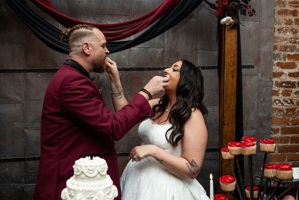 Groom in a burgundy suit feeds wedding cake to his smiling bride in a white gown at a moody, rustic reception with candles and a white cake.