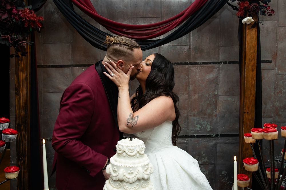 Bride in a white gown and groom in a burgundy velvet suit kiss beside a white tiered wedding cake at a moody, gothic-style reception.