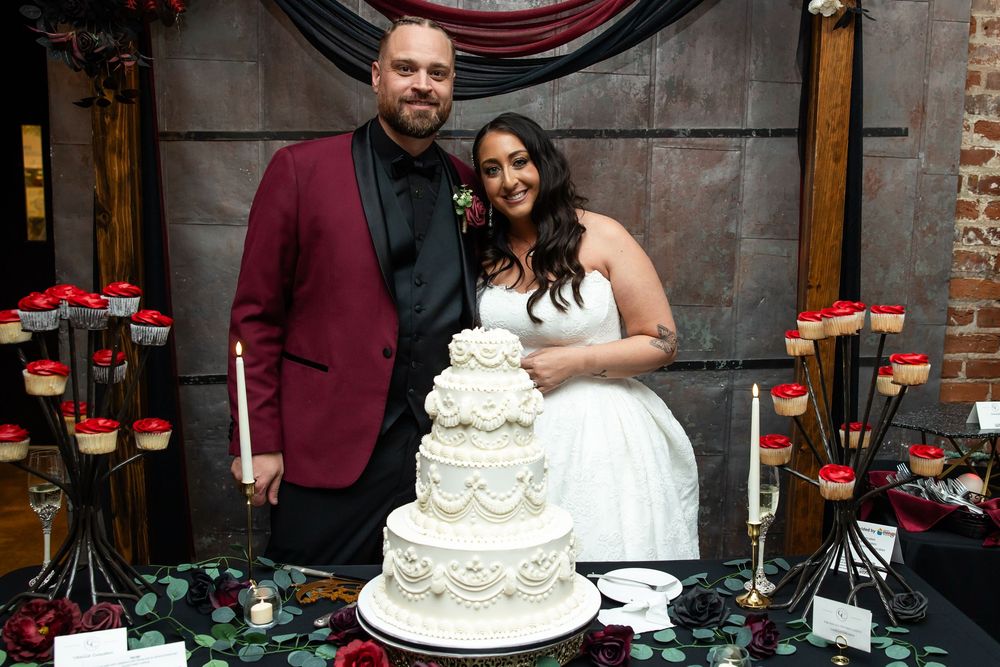 Smiling bride and groom pose beside their elegant white tiered wedding cake surrounded by candles and red dessert stands.
